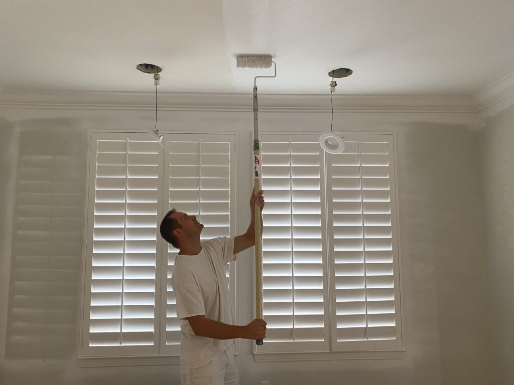 A man in a white T-shirt is painting the ceiling white and pulls out the recessed lights